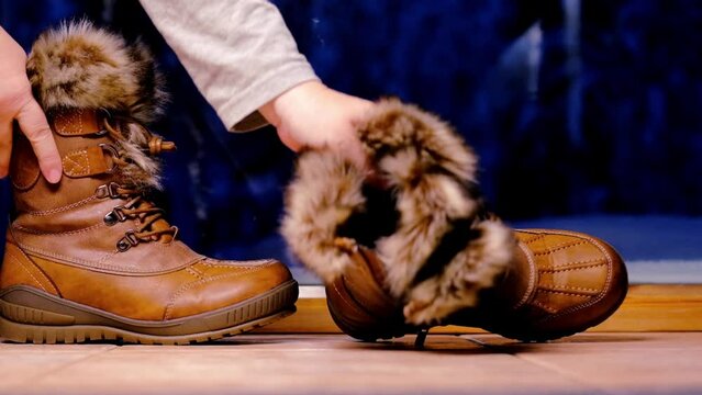 close-up female hands raise pair winter brown boots trimmed with fur from floor, behind large window dark blue evening, concept evening walk, choice warm comfortable seasonal shoes, active recreation