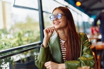 Fototapeta premium Beautiful woman with red hair and glasses sits in the city in a cafe and looks out the window smile with teeth, female freelance blogger