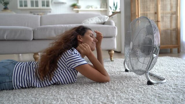 Self-sufficient woman in casual clothes lying on floor in front of running fan enjoying cold wind after going outside. Caucasian girl smiling closing eyes has good time at home in summer weather