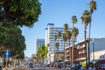 a long street with cars driving and white high rise hotels and office buildings in the city skyline with tall lush green palm trees, people walking and a clear blue sky in Santa Monica California USA