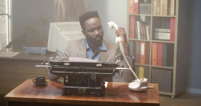 Retro multiracial male working in vintage interior of office. Man sitting at the old typewriter and answering at the phone at the table. Serious businessman of 80's
