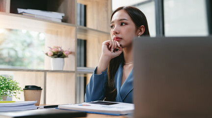 Thoughtful Asian businesswoman sitting at office desk, looking outside, using laptop