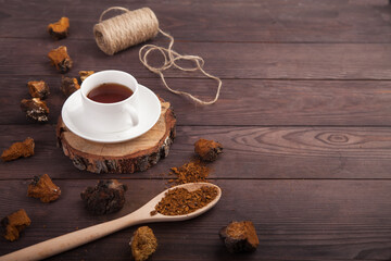 Chaga mushroom coffee drink in a white cup on a wooden stand, scattered pieces of chaga and a spoon with chaga powder on a wooden background. Copy spaes.