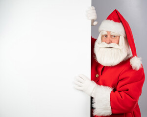 Santa Claus peeks out from behind a white background and points to an empty space with his finger. Merry Christmas. 