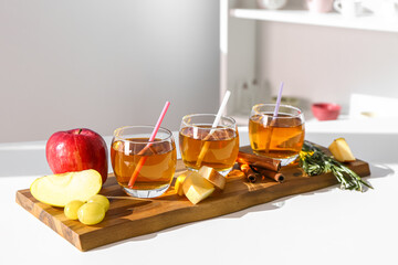 Wooden board with glasses of fresh apple juice, spices fruits on table in kitchen