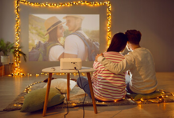View from behind of couple watching photos with video projector. Happy family couple sitting on floor in cosy room decorated with garland lights and hugging in front of projector wall