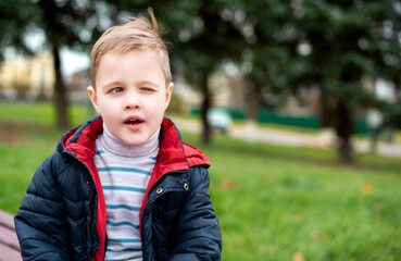 A boy with blond hair winks with one eye. He stands on a background of blurred green trees. The photo is blurred. High quality photo