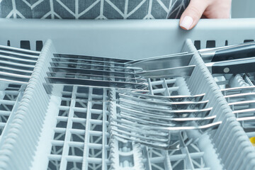 Unrecognizable woman holding grey compartment of dishwasher with metal cutlery set of knives, spoons, forks. Appliance.