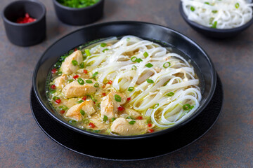 Chicken Rice Noodle Soup. Fried slices of chicken fillet in a yellow broth with diced hot pepper and scallion served in black plate. Selective focus.