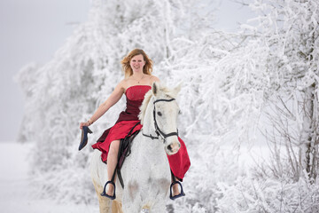 Fototapeta na wymiar junge Frau im roten Kleid reitet auf einem Schimmel in wunderbarer Winterlandschaft mit den Schuhen in der Hand