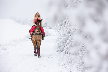 junge Frau im roten Ballkleid reitet durch die Winterlandschaft