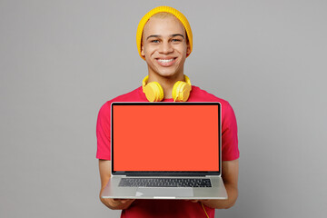 Young IT man of African American ethnicity 20s he wear pink t-shirt yellow hat headphones hold use work on laptop pc computer with blank screen workspace area isolated on plain grey background studio.