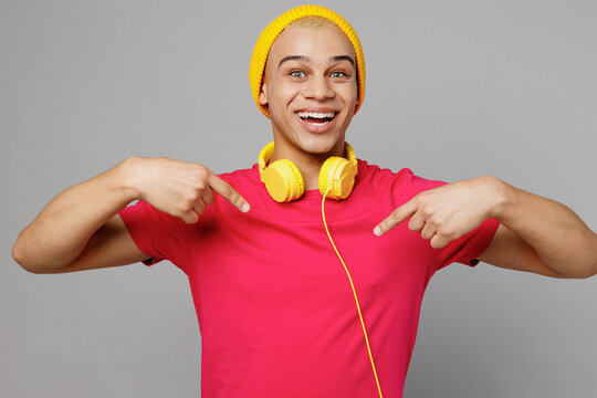 Young confident excited satisfied happy fun cool man of African American ethnicity wear pink t-shirt yellow hat headphones pointing thumb fingers on himself isolated on plain grey background studio