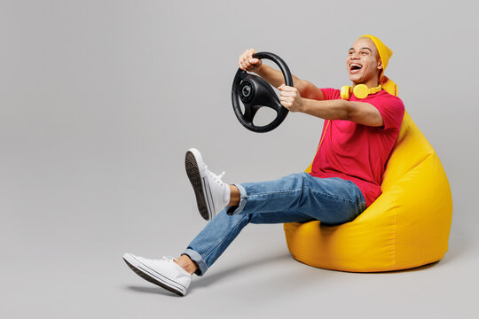 Full Body Happy Young Man Of African American Ethnicity 20s He Wear Pink T-shirt Yellow Hat Headphones Sit In Bag Chair Hold Steering Wheel Driving Car Play Isolated On Plain Grey Background Studio.