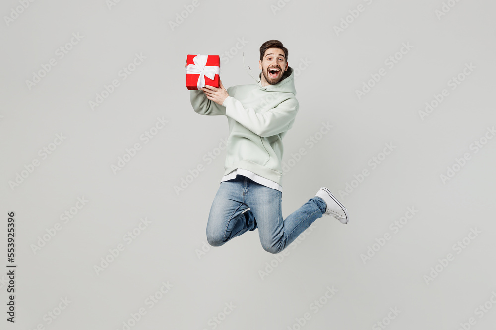 Wall mural Full body surprised amazed young caucasian man wear mint hoody jump high hold red present box with gift ribbon bow isolated on plain solid white background studio portrait. People lifestyle concept.