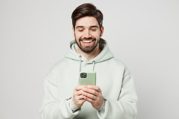 Young smiling cheerful happy caucasian man wear mint hoody hold in hand use mobile cell phone in...