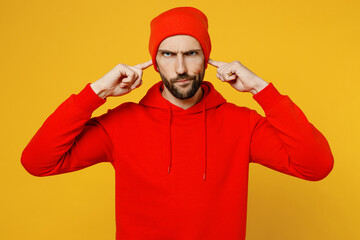 Young sad caucasian man wear red hoody hat look camera cover ears with hands fingers do not want to listen scream isolated on plain yellow color background studio portrait. People lifestyle concept.