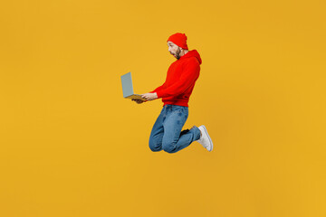Fototapeta na wymiar Full body side view programmer surprised young man wear red hoody hat jump high hold use work on laptop pc computer isolated on plain yellow color background studio portrait. People lifestyle concept.