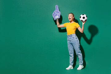 Full body sideways happy young woman fan wear yellow t-shirt foam 1 glove finger up cheer up support football sport team hold soccer ball watch tv live stream isolated on dark green background studio.