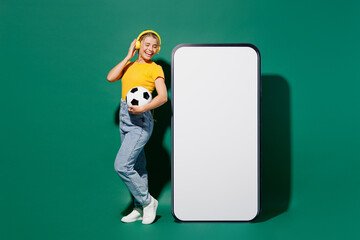Full body young woman fan in t-shirt heaphones listen music cheer up support football sport team hold soccer ball watch tv live stream big blank screen mobile cell phone isolated on green background.