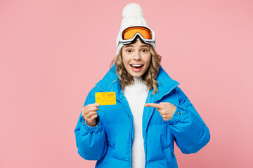 Snowboarder woman wear blue suit goggles mask hat ski padded jacket hold point finger on of credit bank card isolated on plain pink background. Winter extreme sport hobby weekend trip relax concept.