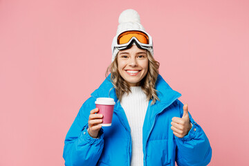 Snowboarder woman wear blue suit goggles mask hat ski padded jacket hold paper cup coffee to go show thumb up isolated on plain pastel pink background. Winter extreme sport hobby trip relax concept.