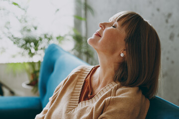 Close up side view tranquil fun elderly woman 50s years old wears casual clothes sits on blue sofa close eyes dream stay at home flat rest relax spend free spare time in living room indoor grey wall.