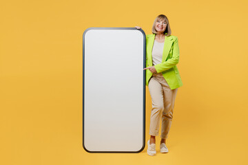 Full body elderly woman 50s years old wear green jacket white t-shirt stand near big huge blank screen mobile cell phone show smartphone with area isolated on plain yellow background studio portrait.
