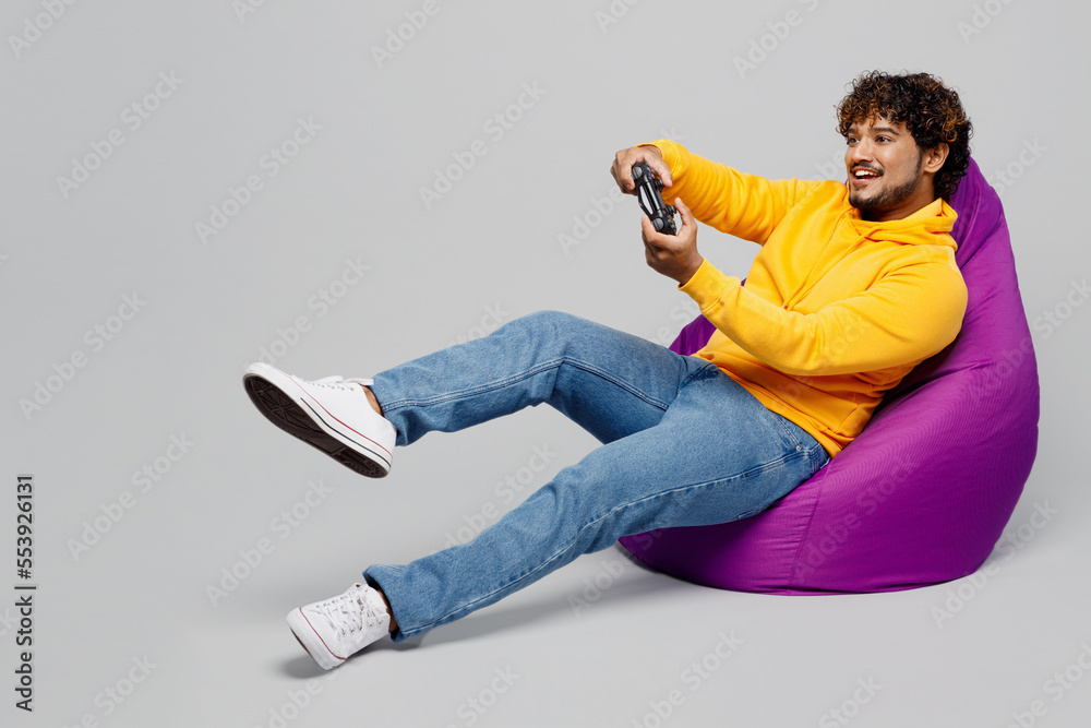 Wall mural Full body young Indian man he wearing casual yellow hoody sit in bag chair hold in hand play pc game with joystick console isolated on plain grey background studio portrait. People lifestyle portrait.