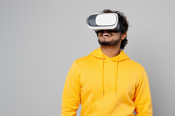 Young gambling fun cool happy smiling Indian man 20s he wearing casual yellow hoody watching in vr headset pc gadget isolated on plain grey color background studio portrait. People lifestyle portrait.