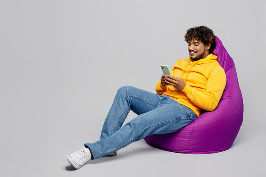 Full body smiling young Indian man 20s he wearing casual yellow hoody sit in bag chair hold in hand use mobile cell phone isolated on plain grey background studio portrait. People lifestyle portrait.
