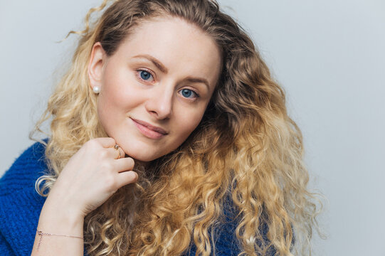 Headshot of adorable blue eyed female model with curly hair, blue eyes, holds chin, looks with pleased expression as listens interlocutor with great interest, isolated over blue studio background