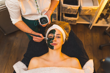medium closeup top view of a woman with a cosmetic facial mask lying on a bed at a spa and a cosmetologist putting more mask, beauty concept. High quality photo