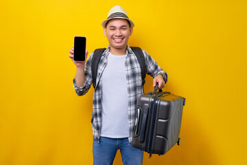 Cheerful young traveler tourist asian man in casual clothes hat holding suitcase and showing blank...
