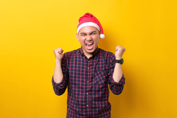 Excited young Asian man in Santa hat raising hands up, celebrating success on yellow studio background. Happy New Year 2023 celebration merry holiday concept