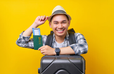 Smiling tourist Asian man in casual clothes hat and backpack holding passport ticket, leaning on suitcase on yellow background. Passenger travel abroad weekends getaway Air flight journey concept