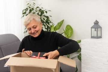 Positive middle aged older woman sitting on couch with carton box on laps, feeling curious about...