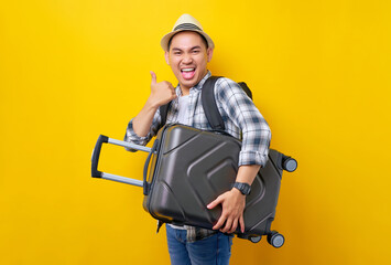 Excited traveler tourist Asian man in casual clothes hat backpack holding suitcase bag, shows thumb up gesture on yellow background. Passenger travel abroad weekends getaway Air flight journey concept