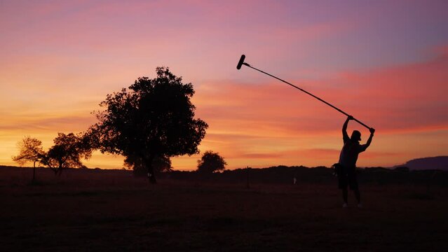 Silhouette nature field and colorful sky on sunset, The boom operate is holding up high microphone boom on set, a part of recording team in film crew production  