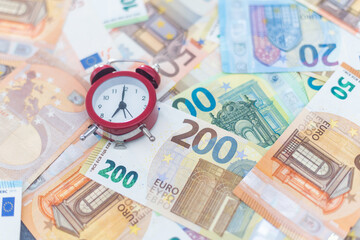 The clock stands on the euro banknotes, next to the coins. The concept is time to make money, every minute bears money losses, every minute can be earned, time is worth money. Close-up.