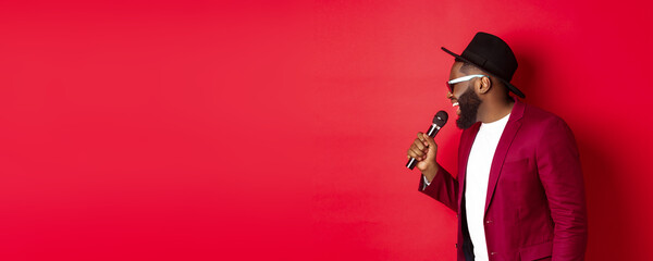 Passionate black male singer performing against red background, singing into microphone, wearing party outfit, standing over red background - obrazy, fototapety, plakaty