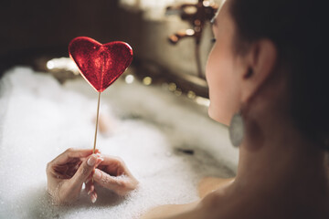 beautiful sexy girl in a bubble bath holds a heart-shaped sweet candy on a stick in her hands