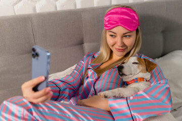 Portrait of beautiful joyful woman in pajamas hugging dog using mobile phone for selfie smiling...