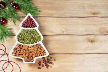 A tree-shaped vase filled with three kinds of seeds and nuts, including red peanuts, green pumpkin seeds, and brown almonds. View from above. On a wooden background with the addition of spruce branche