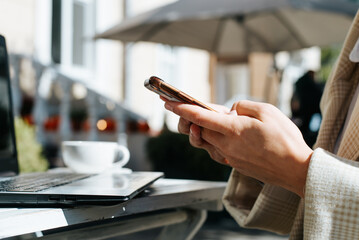 Business woman freelancer holding mobile phone working at table with laptop outdoors. Female hands using smartphone in street cafe. Close-up, side view