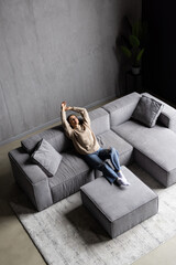 Full length peaceful woman with closed eyes resting on couch, leaning back with hands behind head,...