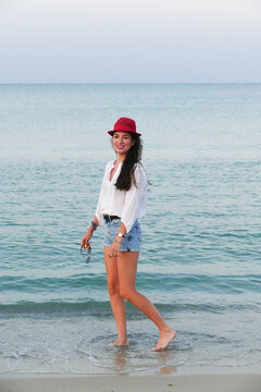 Slim girl walking by the sea. Summer lifestyle image of happy woman walking on the beach. Pretty young woman with long hair  in a white shirt and denim shorts.