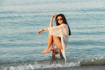A young beautiful woman poses on the ocean beach. Pretty brunette in white beach tunic and black glasses sitting on a stool on the sea.  