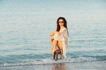 Fototapeta na wymiar A young beautiful woman poses on the ocean beach. Pretty brunette in white beach tunic and black glasses sitting on a stool on the sea. 