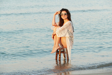 Fototapeta na wymiar A young beautiful woman poses on the ocean beach. Pretty brunette in white beach tunic and black glasses sitting on a stool on the sea. 